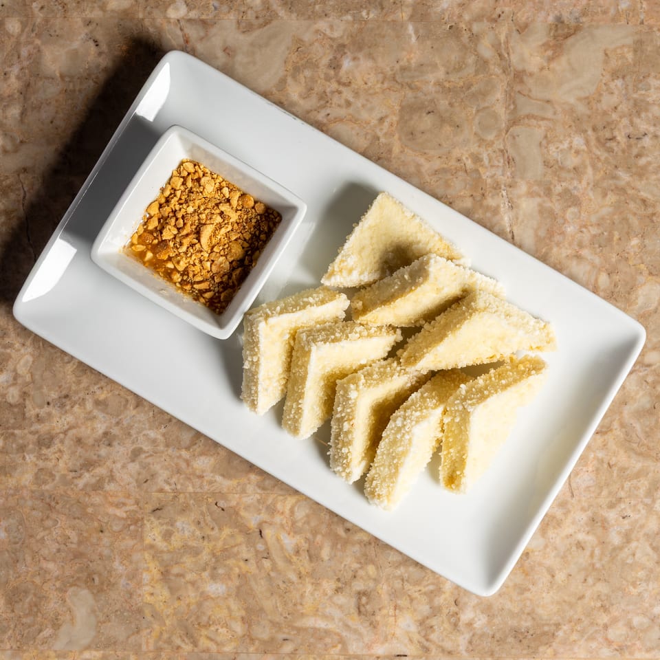 A white rectangular plate holds triangular pastries and a small bowl of crushed peanuts.