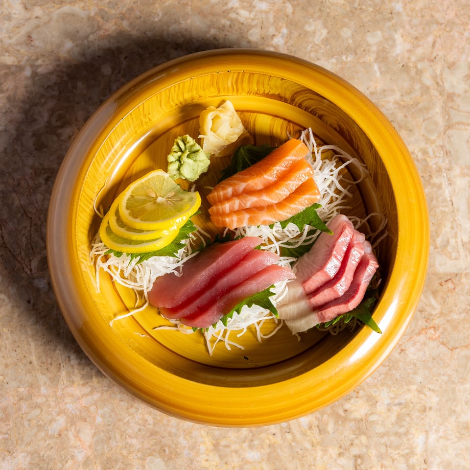 Assorted sashimi slices, including salmon and tuna, with lemon, wasabi, and ginger in a yellow bowl.