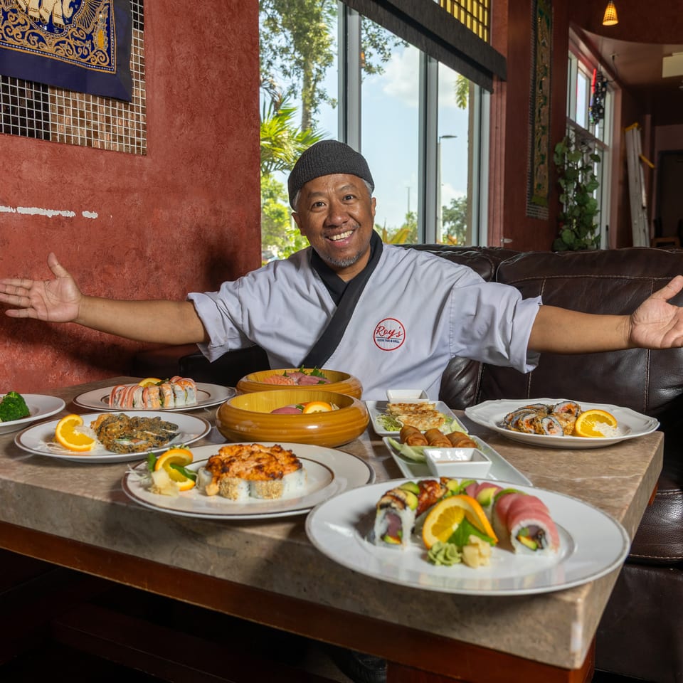 Chef Roy smiles, presenting a table laden with various sushi rolls, sashimi, and other dishes.