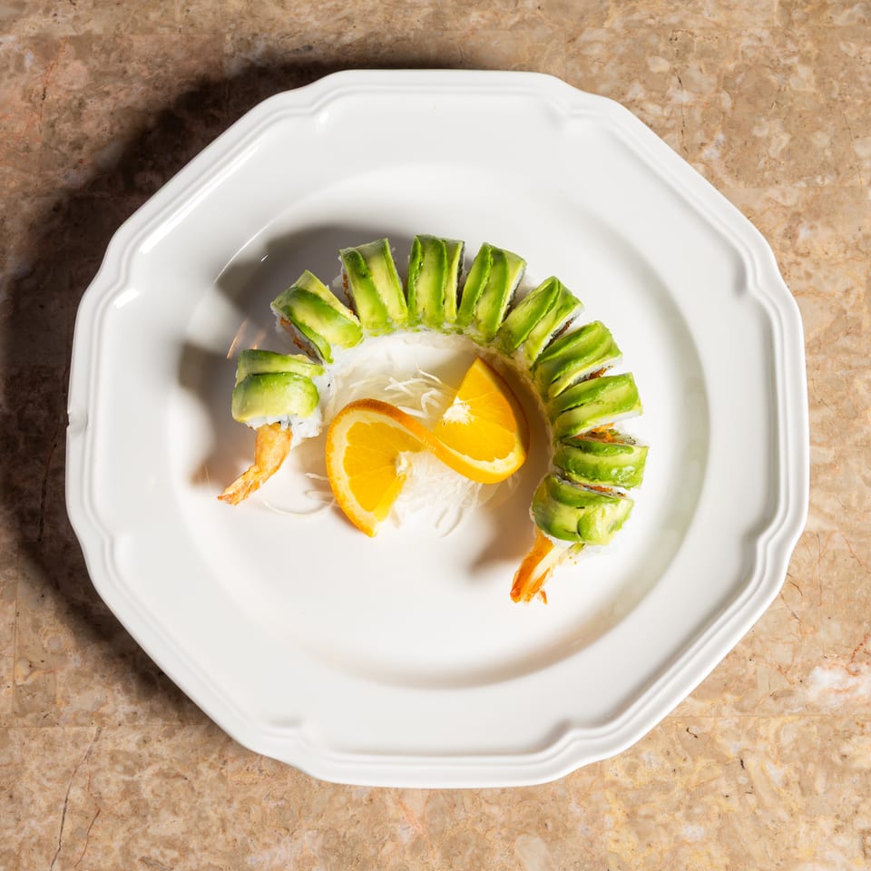 Dragon roll sushi, with avocado slices and shrimp tails, garnished with orange slices on a white plate.