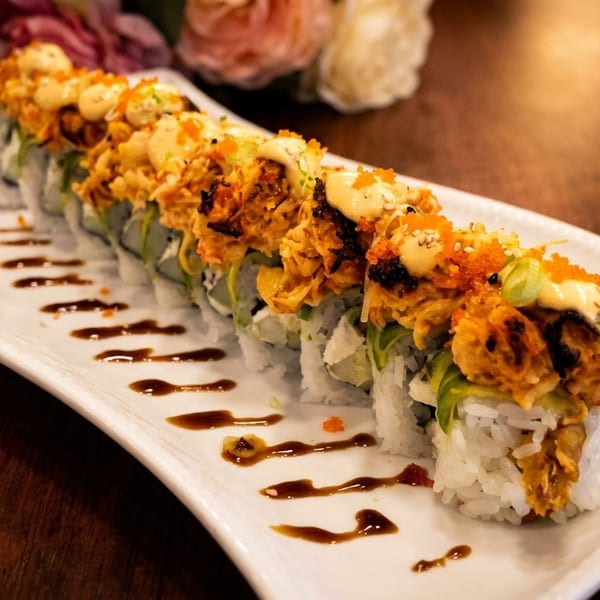 Long sushi roll topped with baked crab, creamy sauce, orange tobiko, and green onions, on a white plate with dark drizzles.
