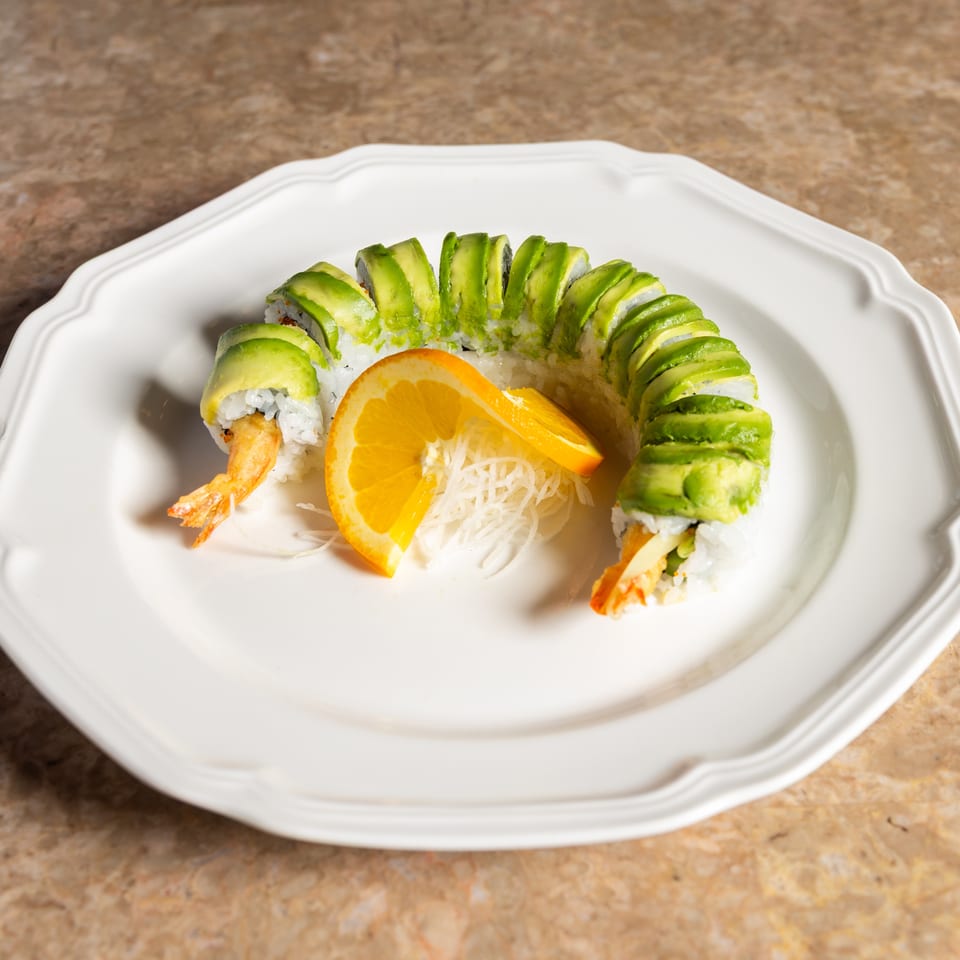 Dragon roll sushi with avocado slices, tempura shrimp, and an orange slice on a white plate.